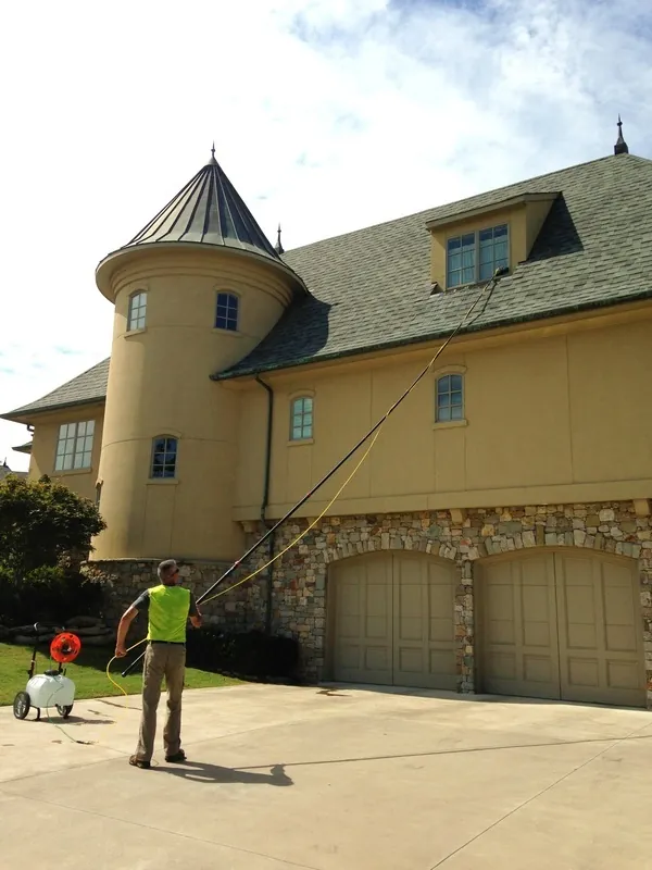 A window cleaner in a safety vest using a water-fed pole on a stucco home with a round tower.
