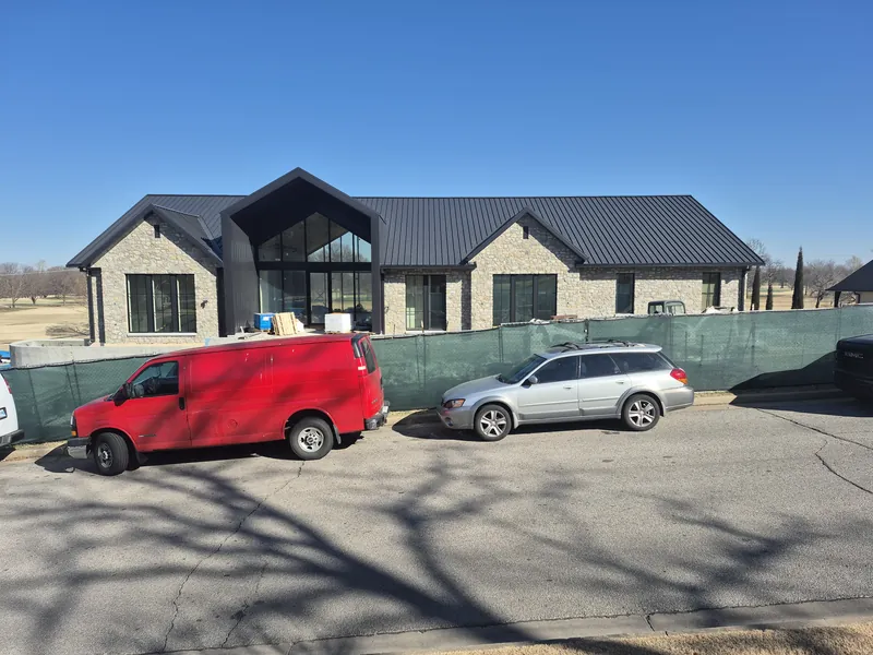 A new stone home with a black metal roof and a glass gable at the center, behind a construction fence.