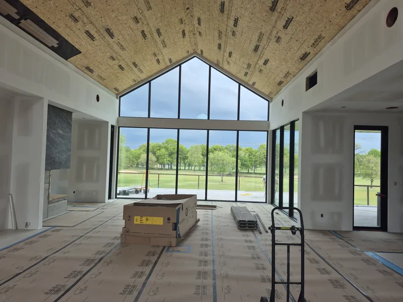 A great room under construction with a vaulted ceiling and a full gable wall of glass facing a green lawn.