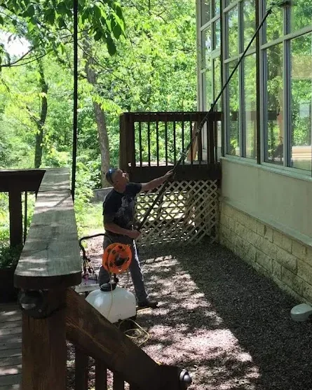 A window cleaner on a shaded gravel path, working a water-fed pole up a tall wall of windows.