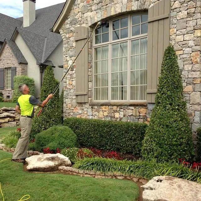 A window cleaner in a safety vest using a water-fed pole on the tall front window of a stone home.