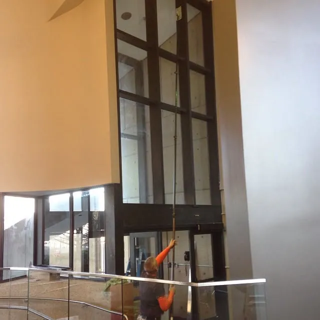 A window cleaner using a pole on tall lobby windows from inside a building.