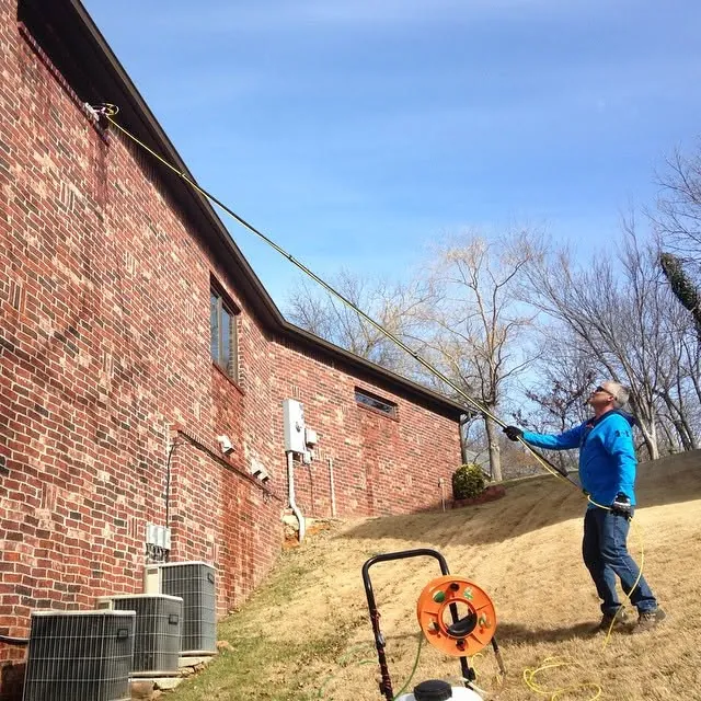 A window cleaner with a water-fed pole along the side of a brick building, with the hose reel on the grass.