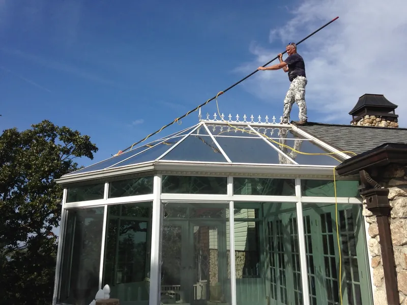 A window cleaner standing on a roof, cleaning a glass conservatory with a long pole.